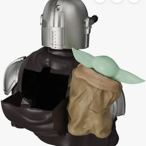 K&ouml;nnte beinhalten: Eine silberfarbene Figur mit Helm, dunkelbraunem Umhang und einer kleinen gr&uuml;nen Kreatur mit gro&szlig;en Ohren in einer beigen Tasche. Die Figur hat einen hohlen R&uuml;ckenbereich mit einem offenen Fach, metallische Schulterpanzer und ein Lautsprechergitter.