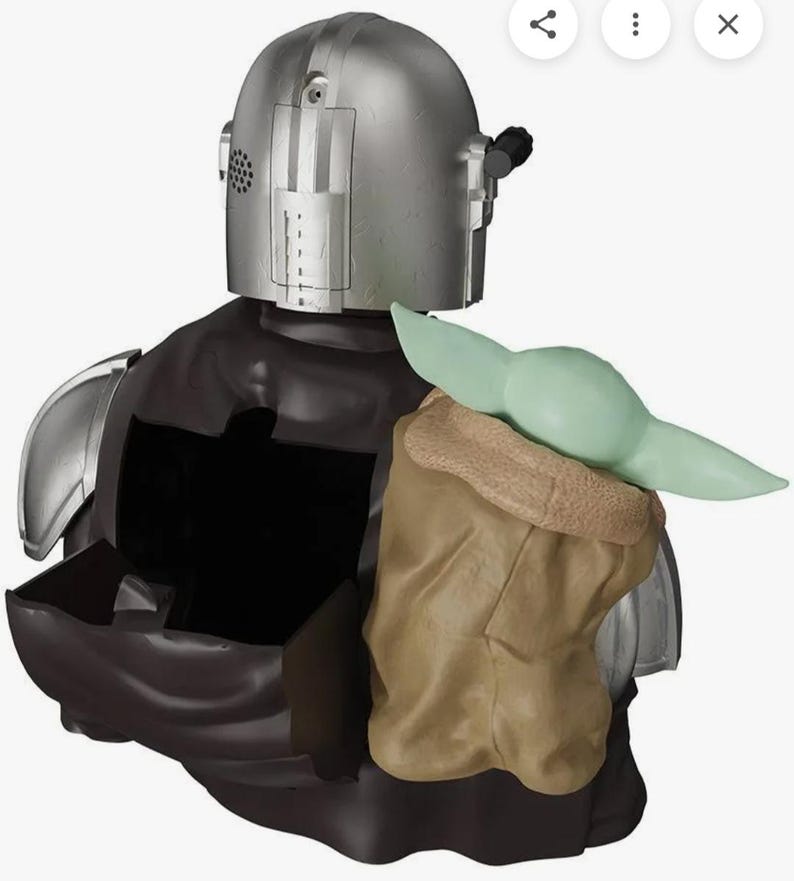 K&ouml;nnte beinhalten: Eine silberfarbene Figur mit Helm, dunkelbraunem Umhang und einer kleinen gr&uuml;nen Kreatur mit gro&szlig;en Ohren in einer beigen Tasche. Die Figur hat einen hohlen R&uuml;ckenbereich mit einem offenen Fach, metallische Schulterpanzer und ein Lautsprechergitter.