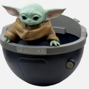 May include: A small green creature with large ears and black eyes, wearing a tan textured robe, sits inside a dark blue, bowl-shaped pod. The pod features a gray rim with rectangular detailing and a small circular button on the side of the exterior.
