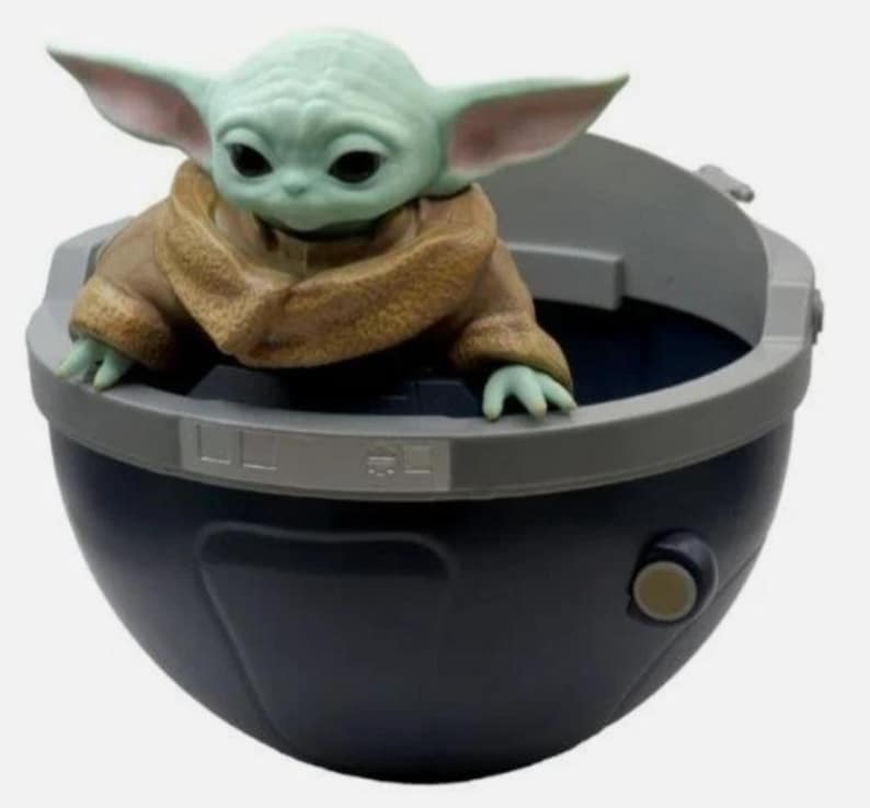 May include: A small green creature with large ears and black eyes, wearing a tan textured robe, sits inside a dark blue, bowl-shaped pod. The pod features a gray rim with rectangular detailing and a small circular button on the side of the exterior.