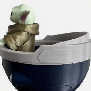 May include: A small green creature with large ears and a tan robe sits inside a dark blue, bowl-shaped container. The container features a wide, light gray rim that curves upward, creating a distinct border around the top edge of the object.
