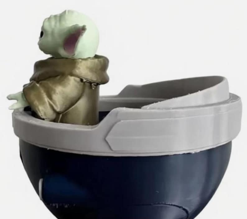 May include: A small green creature with large ears and a tan robe sits inside a dark blue, bowl-shaped container. The container features a wide, light gray rim that curves upward, creating a distinct border around the top edge of the object.