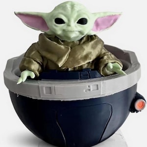 May include: A small green creature with large ears and black eyes sits inside a dark blue, bowl-shaped pod. The creature wears a tan-colored robe with a high collar. The pod features a light gray rim and a small orange circular detail on the side.