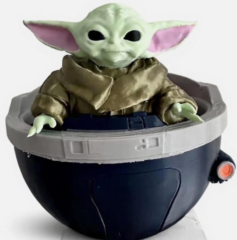 May include: A small green creature with large ears and black eyes sits inside a dark blue, bowl-shaped pod. The creature wears a tan-colored robe with a high collar. The pod features a light gray rim and a small orange circular detail on the side.