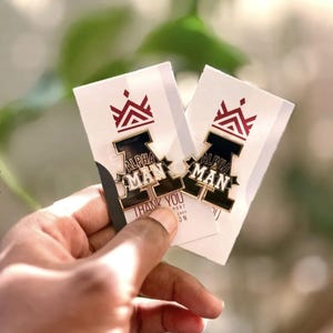 May include: Two enamel pins with the text "ALPHA MAN" in white lettering on a black background, with a gold border. Each pin is on a white card with a red and white crown design and the words "THANK YOU" at the bottom.