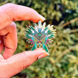 May include: A gold-colored enamel pin with a green, blue, and white design. The pin features a stylized face with feathers and a dripping effect.