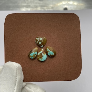 May include: Four polished turquoise gemstones with brown matrix, including a heart shape and three teardrop shapes, are displayed on a brown card. The gemstones have a unique pattern of blue and brown colors. The gemstones are set against a white background.