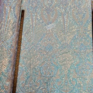 May include: A close-up of iridescent blue and white sequin fabric with a damask pattern. The fabric is measured with a ruler in inches.