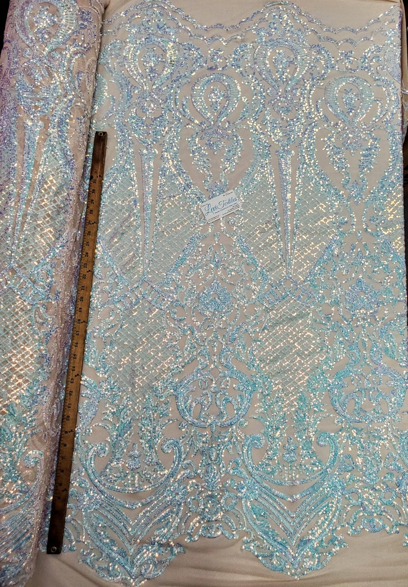 May include: A close-up of iridescent blue and white sequin fabric with a damask pattern. The fabric is measured with a ruler in inches.