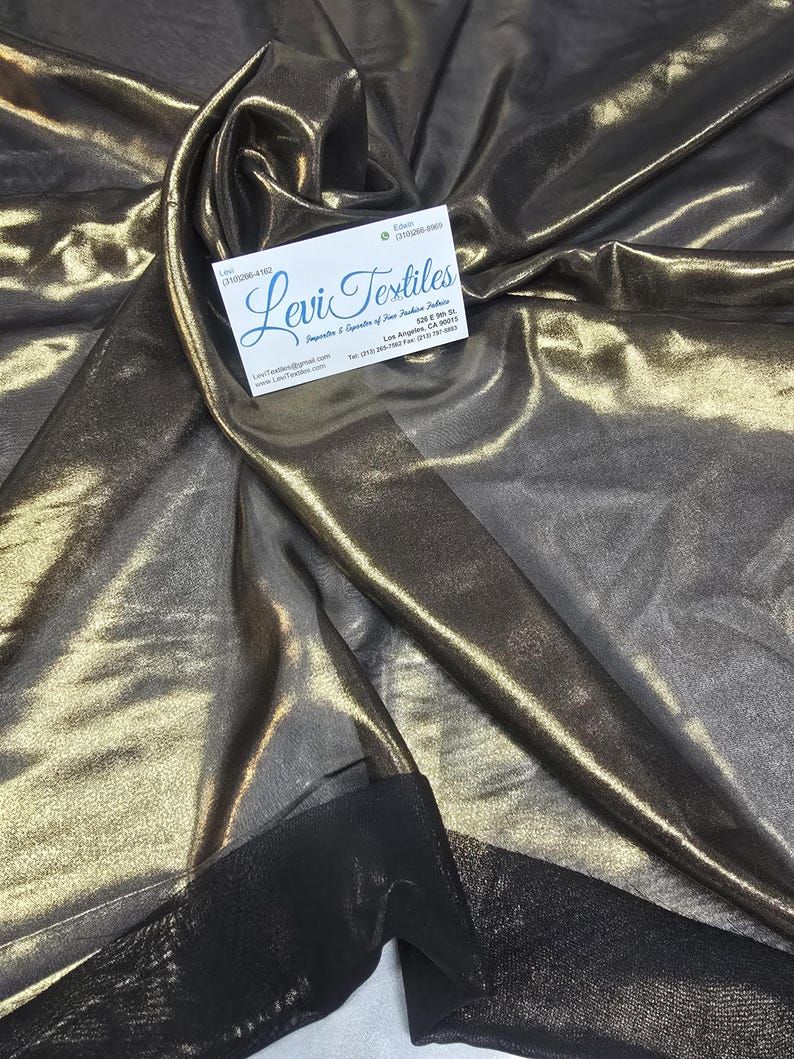 May include: Metallic fabric in shades of gold and bronze, with a shimmering, reflective quality. The fabric is draped, creating folds and shadows. A business card with the text "Levi Textiles" is visible.