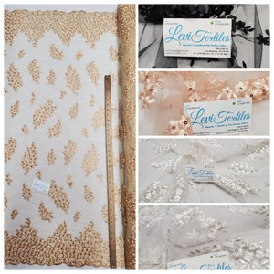 May include: A roll of beige embroidered lace fabric with a floral pattern. The fabric is shown with a measuring tape and a business card for Levi Textiles.