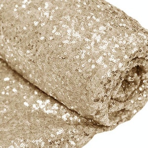 Könnte beinhalten: Eine Rolle schimmernden Stoffes, bedeckt mit kleinen, runden, champagnerfarbenen Pailletten. Der Stoff ist gefaltet, wodurch die Textur und der Glanz der Pailletten sichtbar werden. Der Gesamteindruck ist luxuriös und glamourös, geeignet für besondere Anlässe oder Dekorationsprojekte.