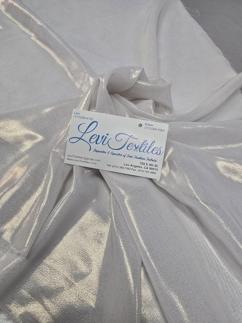 May include: White, shimmering fabric with soft folds. A business card with the text "Levi Textiles" is placed on the fabric. The fabric appears to be a lightweight material, possibly for fashion or design.