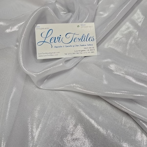 May include: White, shimmering fabric with a soft, flowing texture. The fabric is draped and folded, creating a sense of movement. A business card with the text "Levi Textiles" is placed on top of the fabric.