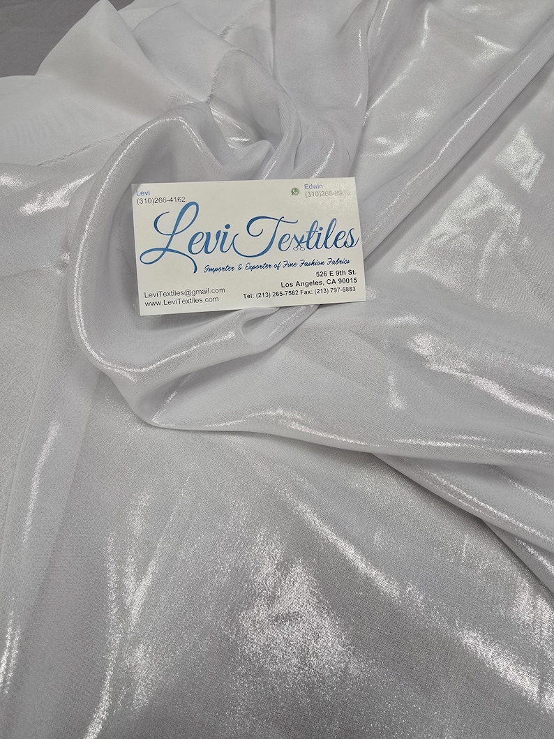 May include: White, shimmering fabric with a soft, flowing texture. The fabric is draped and folded, creating a sense of movement. A business card with the text "Levi Textiles" is placed on top of the fabric.