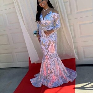 May include: A woman wearing a white sequined gown with a plunging neckline and long sleeves. The gown has a mermaid silhouette and is adorned with floral embroidery. The woman is standing on a red carpet.