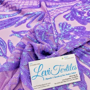May include: Purple iridescent sequins on a sheer lavender fabric. The fabric is draped over a business card that reads "Levi Textiles Importer & Exporter of Fine Fashion Fabrics" with contact information.