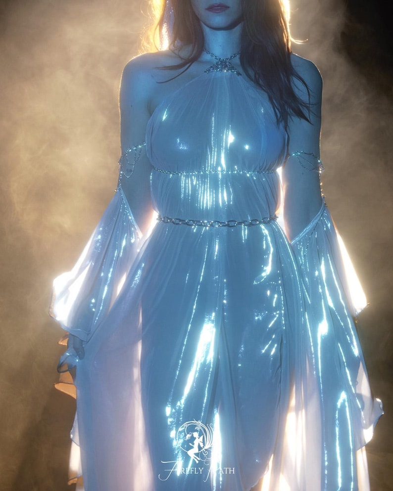 May include: A shimmering, light blue gown with a halter neckline and flowing sleeves. The dress features a silver chain belt and delicate chain accents on the arms. The image is set against a soft, glowing background, with the text "FIREFLY PATH" at the bottom.
