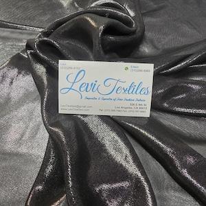 May include: A close-up of a shimmering, metallic gray fabric with soft folds. A business card with the text "Levi Textiles" is visible. The fabric has a subtle sheen, reflecting light and creating a textured appearance. The fabric is suitable for apparel and home decor.