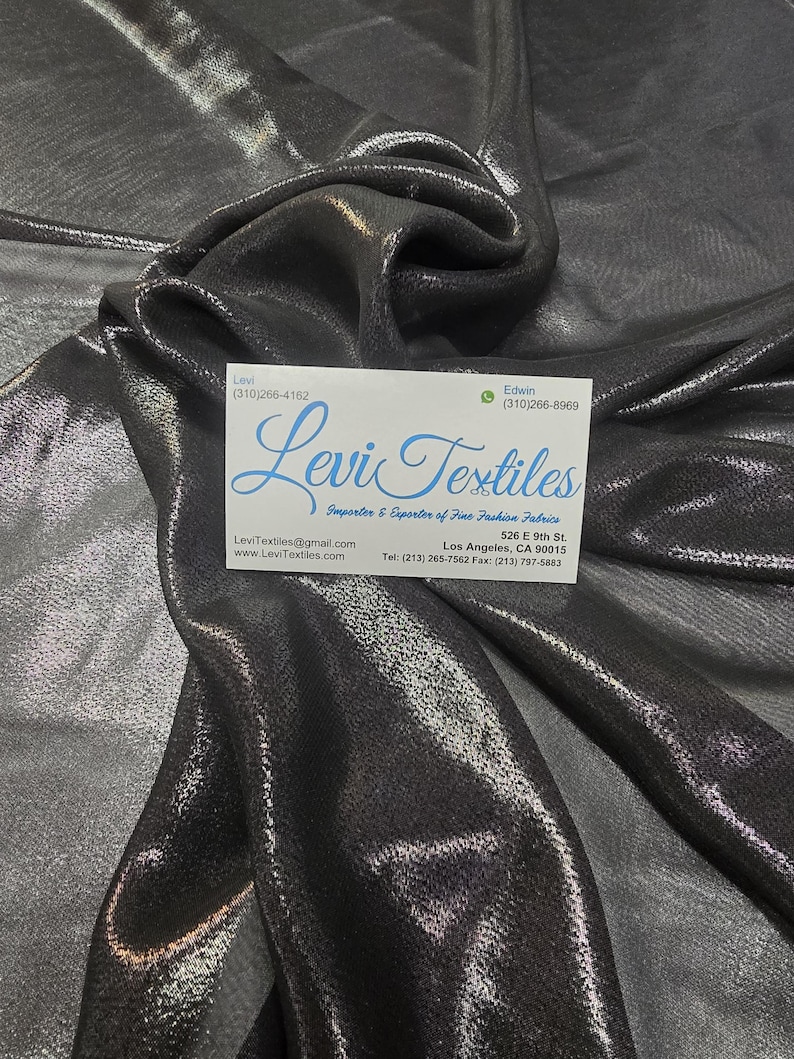 May include: A close-up of a shimmering, metallic gray fabric with soft folds. A business card with the text "Levi Textiles" is visible. The fabric has a subtle sheen, reflecting light and creating a textured appearance. The fabric is suitable for apparel and home decor.