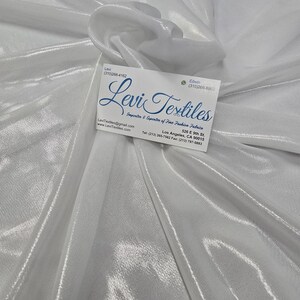 May include: A close-up of a white, shimmering fabric with a subtle texture. A business card with the text "Levi Textiles" is placed on the fabric. The fabric appears to be draped, creating soft folds and highlights.