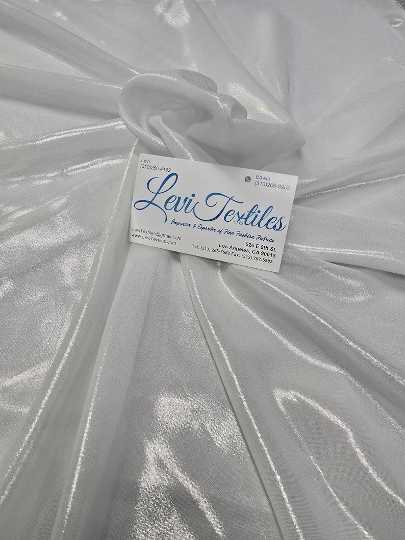 May include: A close-up of a white, shimmering fabric with a subtle texture. A business card with the text "Levi Textiles" is placed on the fabric. The fabric appears to be draped, creating soft folds and highlights.