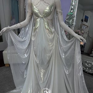 May include: A flowing, ethereal gown with a gold bodice and sheer, white skirt. The dress features delicate silver jewelry and a cape-like overlay. The gown is displayed on a mannequin, showcasing its elegant design and draping.