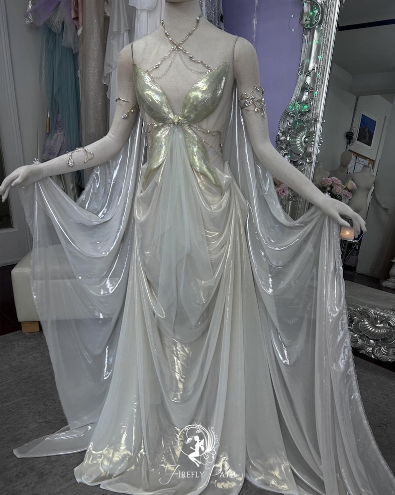 May include: A flowing, ethereal gown with a gold bodice and sheer, white skirt. The dress features delicate silver jewelry and a cape-like overlay. The gown is displayed on a mannequin, showcasing its elegant design and draping.