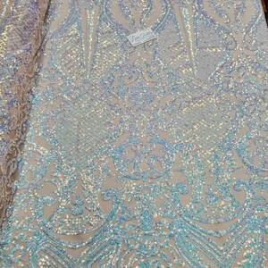 May include: A white iridescent fabric with a blue and white damask pattern. The fabric is covered in sequins and has a glittery finish.