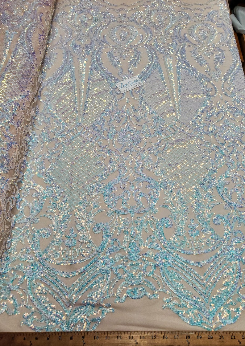 May include: A white iridescent fabric with a blue and white damask pattern. The fabric is covered in sequins and has a glittery finish.
