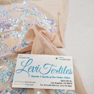 May include: A business card for Levi Textiles, an importer and exporter of fine fashion fabrics. The card includes the company's contact information, including phone numbers, email address, and website. The card is lying on top of a piece of fabric with iridescent sequins.