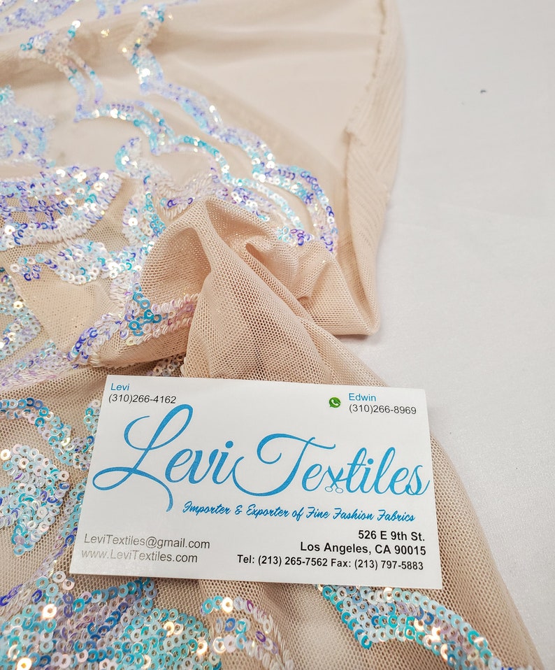 May include: A business card for Levi Textiles, an importer and exporter of fine fashion fabrics. The card includes the company's contact information, including phone numbers, email address, and website. The card is lying on top of a piece of fabric with iridescent sequins.