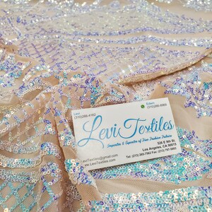 May include: A close-up of a beige sequined fabric with a light blue and purple iridescent sheen. The fabric is draped over a business card with the text "Levi Textiles" and contact information.