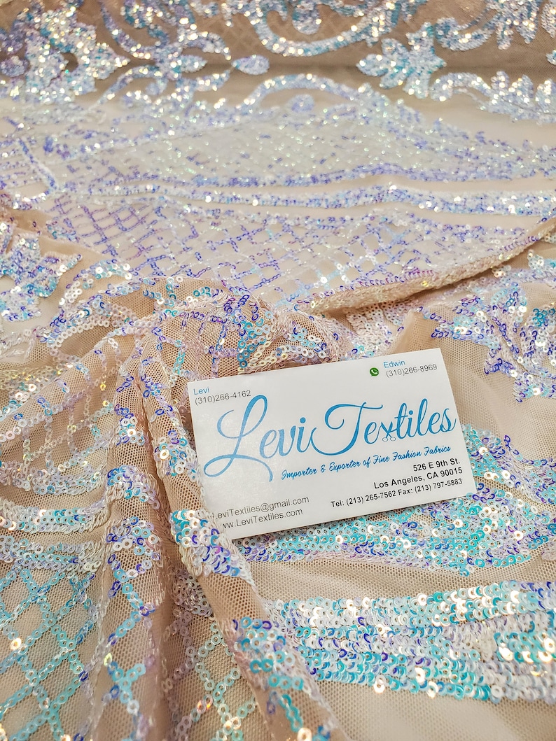 May include: A close-up of a beige sequined fabric with a light blue and purple iridescent sheen. The fabric is draped over a business card with the text "Levi Textiles" and contact information.