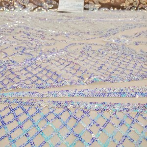 May include: Close-up of iridescent blue, purple, and white sequins sewn onto a beige mesh fabric. The sequins are arranged in a diamond pattern.