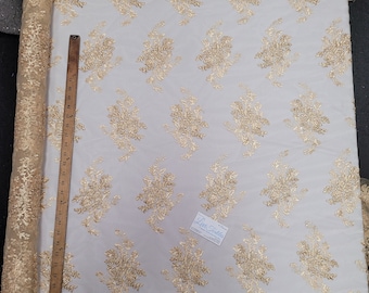 May include: A roll of gold embroidered lace fabric with a floral pattern. The lace has a scalloped edge and is on a white background.