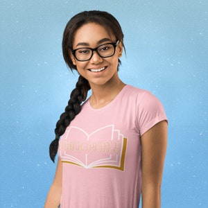 May include: A light pink t-shirt featuring the word "BIBLIOPHILE" in gold lettering above a stylized open book design. The design has white and gold accents. The shirt is a crew neck style.