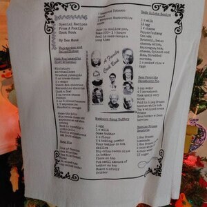 May include: A white tea towel with a printed design featuring a family cookbook, recipes, and a spoon. The recipes include "Grandma's Favorite Chocolate Chip Cookies", "Mother's Sourdough Starter", and "Dad's Favorite Bean Soup".