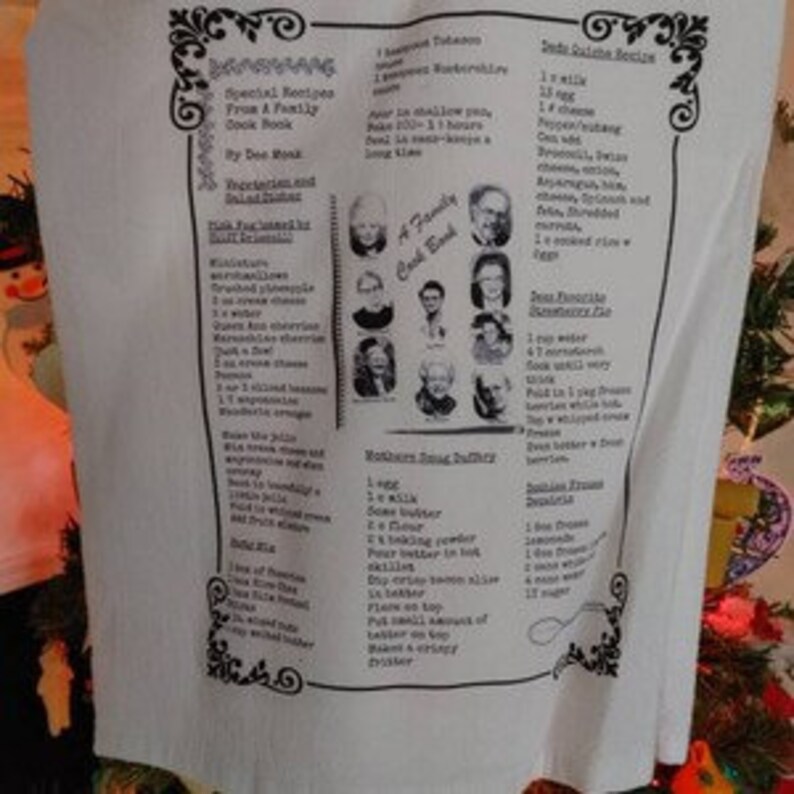 May include: A white tea towel with a printed design featuring a family cookbook, recipes, and a spoon. The recipes include "Grandma's Favorite Chocolate Chip Cookies", "Mother's Sourdough Starter", and "Dad's Favorite Bean Soup".