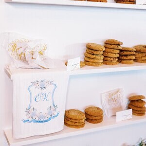 May include: White shelves with a white towel with blue and gold embroidery and the initials "E & K" on it. The towel is on a white box. There are stacks of chocolate chip cookies on the shelves.