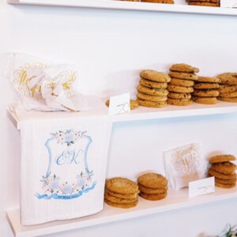 May include: White shelves with a white towel with blue and gold embroidery and the initials "E & K" on it. The towel is on a white box. There are stacks of chocolate chip cookies on the shelves.