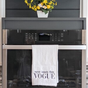 May include: A white kitchen towel with the text "more issues than VOGUE" hanging on a stainless steel oven door. A bouquet of yellow flowers in a white pot is on a shelf above the oven.