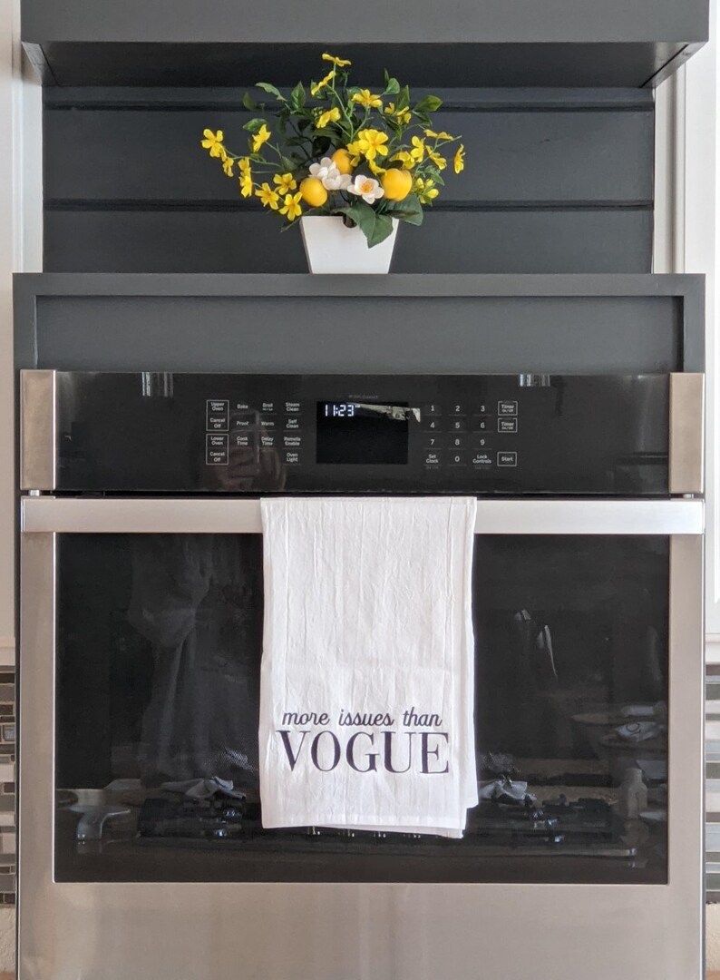 May include: A white kitchen towel with the text "more issues than VOGUE" hanging on a stainless steel oven door. A bouquet of yellow flowers in a white pot is on a shelf above the oven.