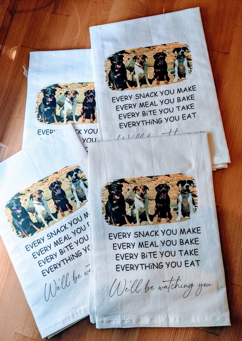 May include: Four dogs are sitting in a row on a white kitchen towel. The towel has the text "Every snack you make, every meal you bake, every bite you take, everything you eat. We'll be watching you." printed on it.