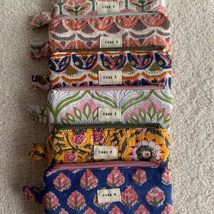 Op de afbeelding: Een stapel van zeven rechthoekige etuis met ritssluiting met diverse bloemen- en paisley-patronen in roze, blauw, geel en groen. Elke etui heeft een klein rechthoekig label met de tekst "EASA T".