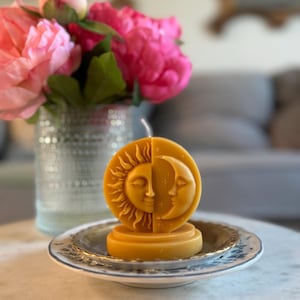 May include: A decorative candle in the shape of the sun and moon, resting on a small, ornate plate. The candle is a warm yellow color, and the plate has a blue and white floral design. A vase of pink flowers is in the background.