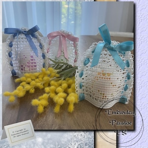 Può includere: Tre sacchetti regalo in crochet bianco con fiocchi di nastro blu, rosa e azzurro. I sacchetti sono pieni di piccoli regali e sono su un tavolo con un mazzo di mimosa gialla. Il testo "Embroidery Paradise ES" è visibile nell'angolo in basso a destra dell'immagine.