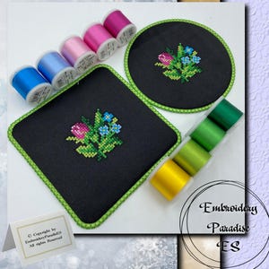May include: Two black embroidery hoops with green trim, one round and one square, with a floral design embroidered in pink, blue, and green. The hoops are surrounded by spools of embroidery floss in various colors.