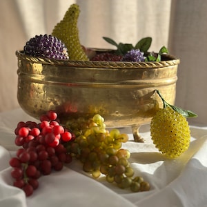 Pode incluir: Uma tigela de latão com uma pêra de contas, uvas e outras frutas de contas. A tigela tem uma borda de corda ao redor da parte superior e fica em três pés. A tigela está sobre uma superfície branca.