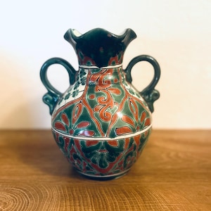 Puede incluir: Un jarrón de cerámica con borde y asas verde oscuro, con un patrón floral rojo y verde. El jarrón tiene un cuerpo bulboso y una parte superior con volantes, sobre un fondo blanco.
